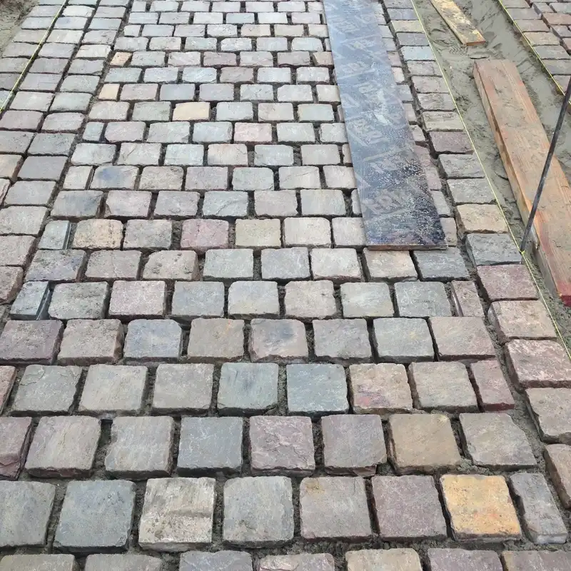 Détail de pavés de porphyre posés en appareil, avec une bande sombre et une règle sur le lit de sable.