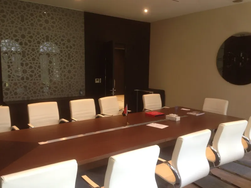 Salle de réunion achevée : longue table en bois, chaises blanches, panneau mural sombre et claustra découpé devant la fenêtre.