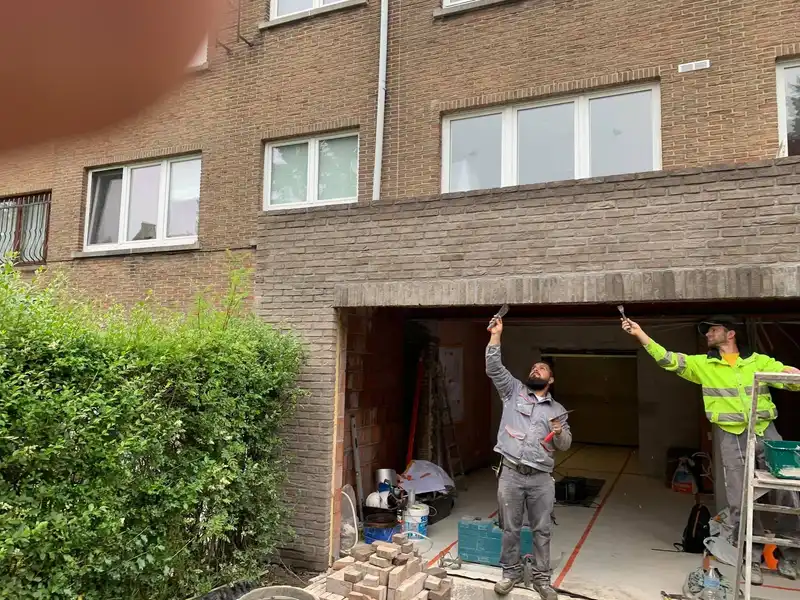 Deux membres de l’équipe maçonnent la brique de parement au-dessus d’une baie de garage ; l’un travaille depuis un escabeau.