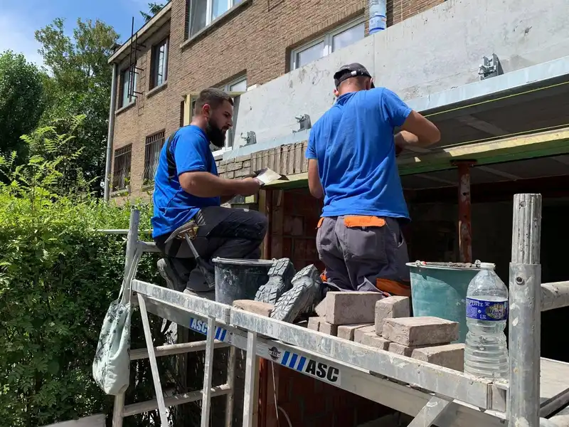 Deux maçons sur un plateau d’échafaudage montent la brique de parement sous un auvent en béton.