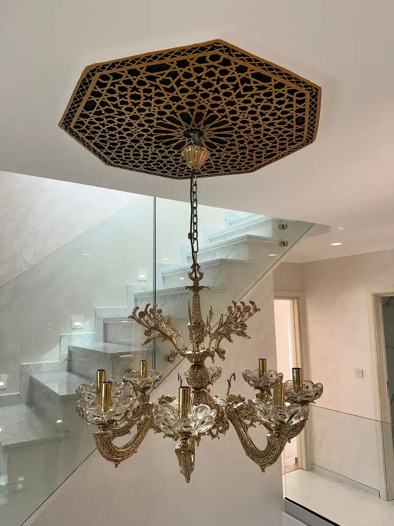 Lustre sous un médaillon de plafond en bois, avec l’escalier en marbre derrière un garde-corps en verre.