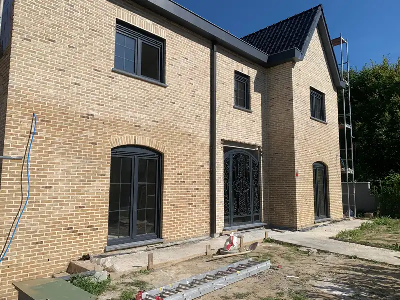 Façade achevée en brique jaune, avec fenêtres cintrées, menuiseries extérieures gris foncé et toiture en tuiles noires à pignon.