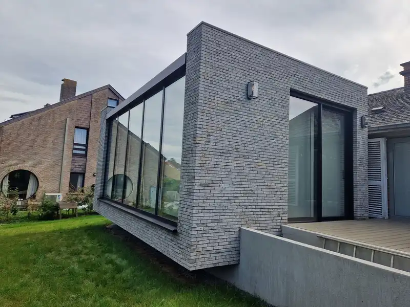 Extension achevée en brique moulée main grise avec une haute paroi vitrée, raccordée à la maison existante, jardin devant.