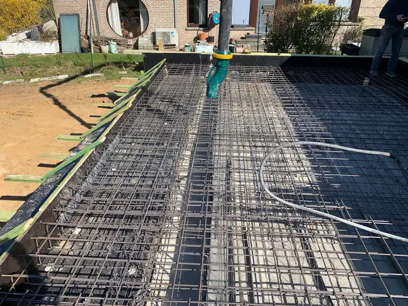 Treillis d’armature sur toute la dalle avec le flexible de la pompe à béton au-dessus, et la maison existante et son œil-de-bœuf à l’arrière.